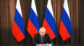 Путин сидит за столом с бумагами в руках на фоне четырех российских флагов